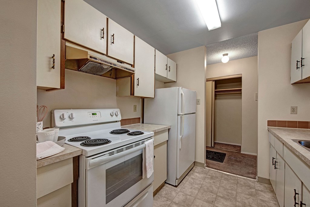 Small, neutral-toned kitchen here at Sterling Place Apartment Homes with white cabinets, electric stove, and refrigerator. Bright lighting and a narrow layout create a cozy atmosphere.