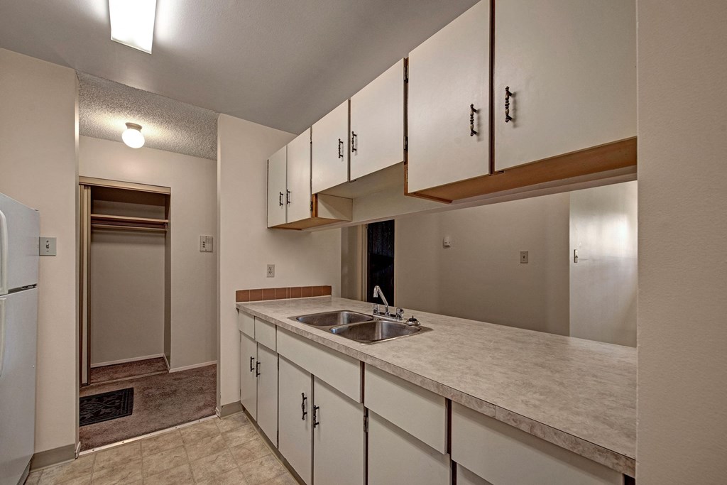 Narrow kitchen here at Sterling Place Apartment Homes with beige cabinets and countertops, a double stainless steel sink, and a fridge. Adjacent room visible through a doorway, creating a cozy feel.