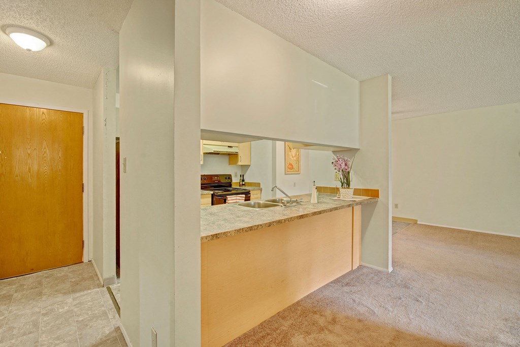 Open kitchen and living area here at Sterling Place Apartment Homes with beige carpet and light tiled flooring. Wooden door on the left, marble countertop with sink, stove, and pink flowers.