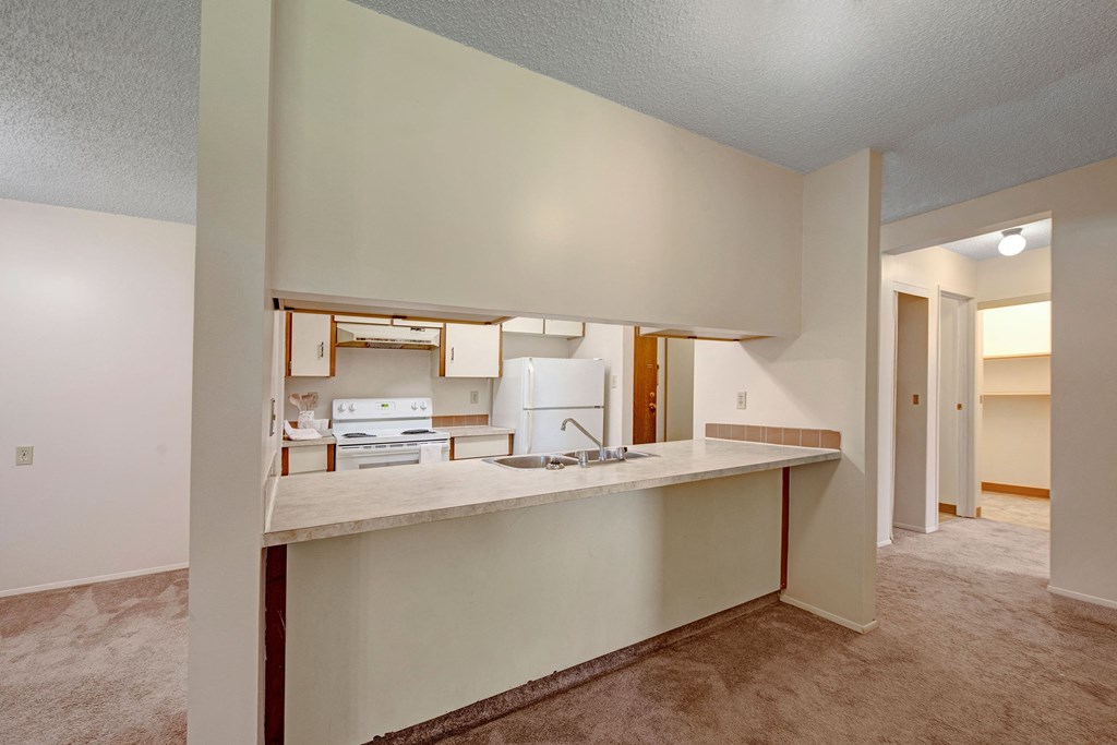 Small, unfurnished kitchen here at Sterling Place Apartment Homes with beige walls and carpet. Features a countertop with sink, white appliances, and an open layout, creating a simple, cozy vibe.