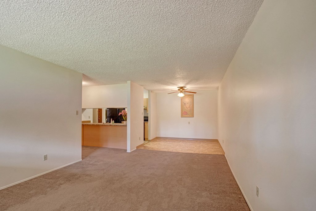 Empty living room here at Sterling Place Apartment Homes with beige carpeting and white walls, leading to an open kitchen area with a ceiling fan. Soft lighting and minimal decor give a neutral, calm feel.