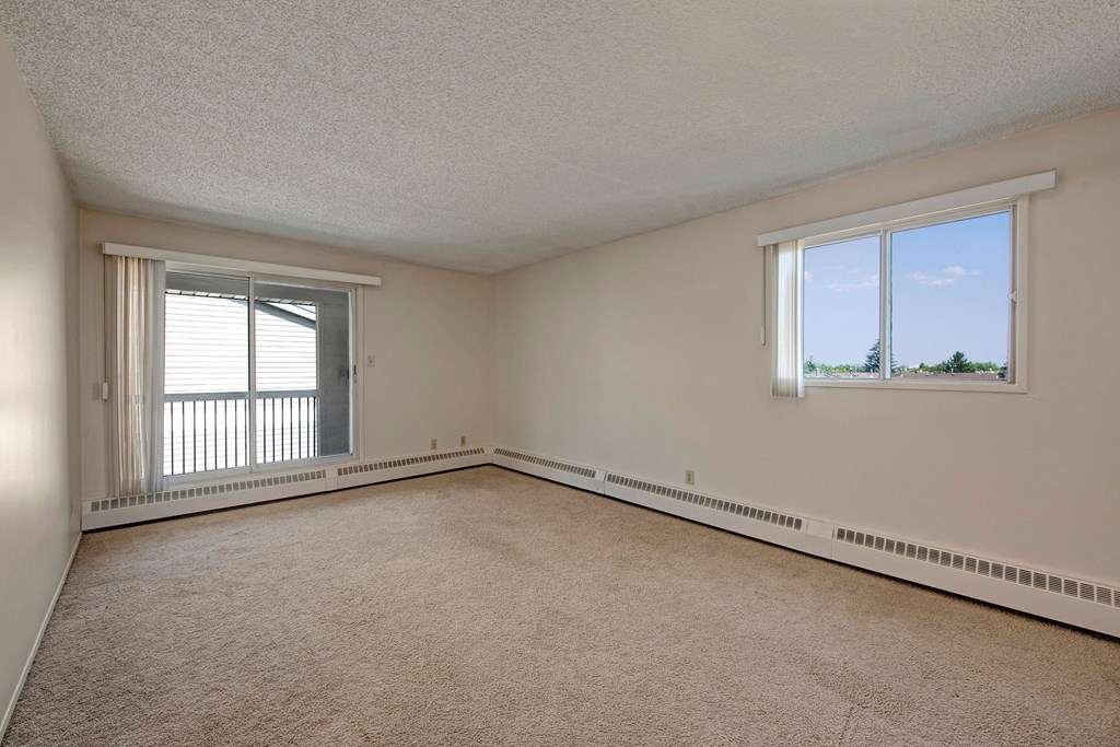 A spacious, empty room here at Sterling Place Apartment Homes with beige carpet and white walls. A large sliding glass door and a window let in natural light, offering views of trees outside.