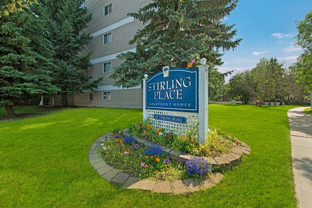 Sign for Stirling Place Apartment Homes, surrounded by colorful flowers and lush grass, with a tall building and evergreen trees in the background.