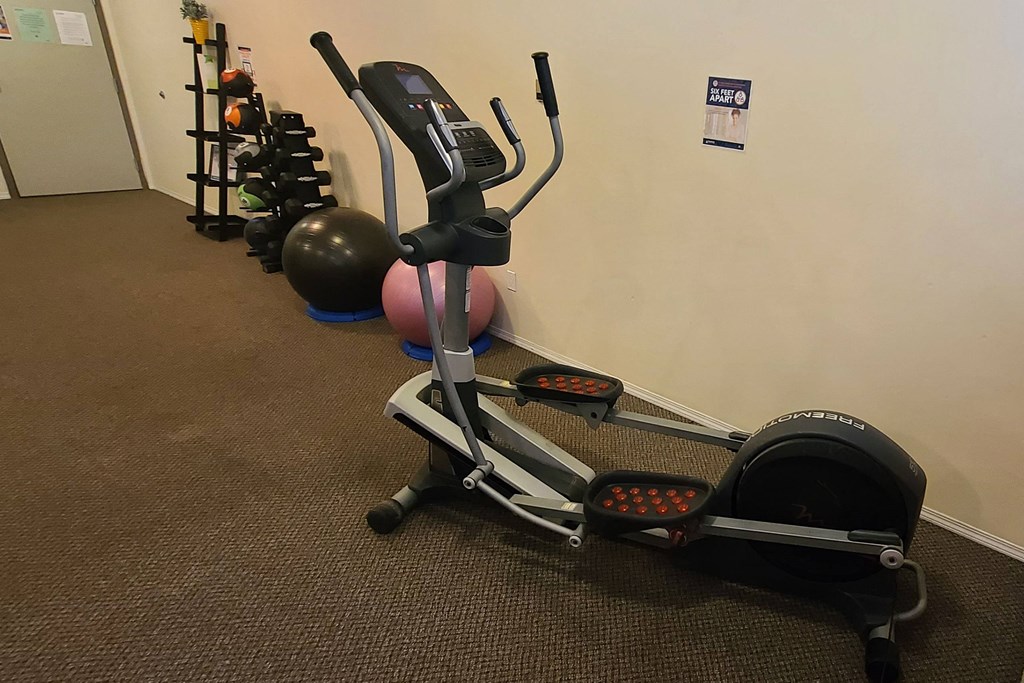 A home gym corner here at Ventana Apartment Homes with an elliptical machine on carpeted floor, exercise balls, and a dumbbell rack. A "six feet apart" sign is on the wall.