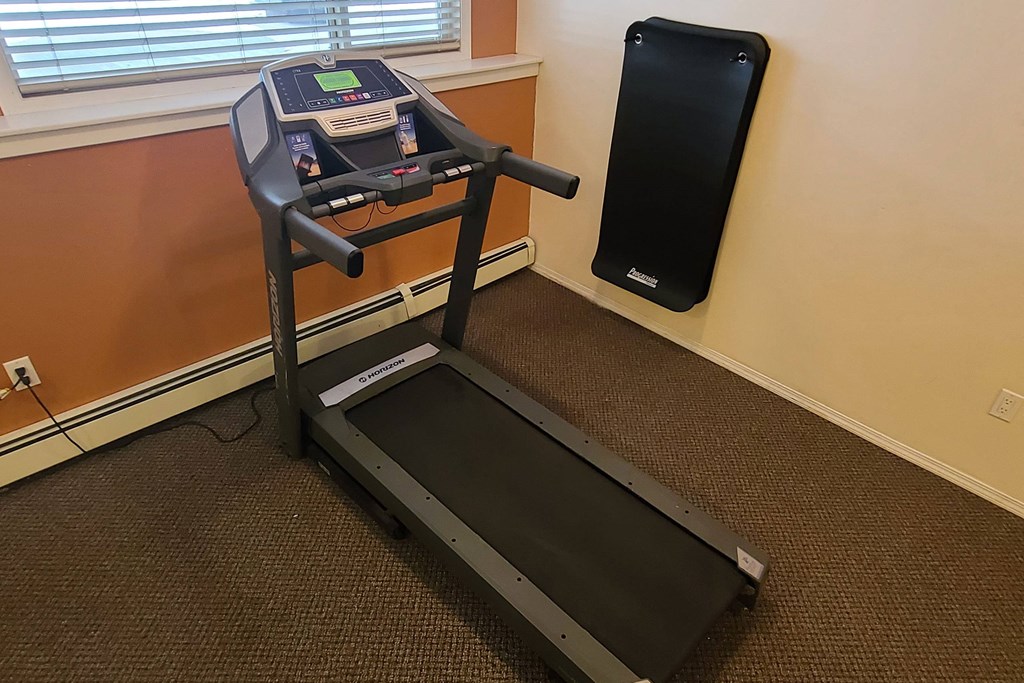 A treadmill in a gym here at Ventana Apartment Homes with orange and beige walls, tan carpet, and a window with blinds. A fitness mat is mounted on the wall beside it.