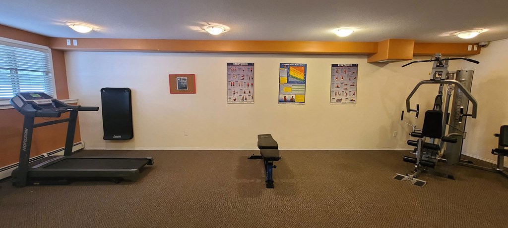 A small gym here at Ventana Apartment Homes with a treadmill on the left, an exercise bench in the center, and a weight machine on the right. Posters on the wall show exercise and heart rate information. The room has an orange and beige color scheme with carpeted flooring, creating a practical and calm atmosphere.