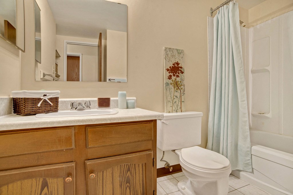 A cozy bathroom here at Ventana Apartment Homes with cream walls, featuring a wooden vanity, white countertop, and wicker basket. A floral artwork hangs above the toilet beside a shower with a light blue curtain, creating a calm atmosphere.
