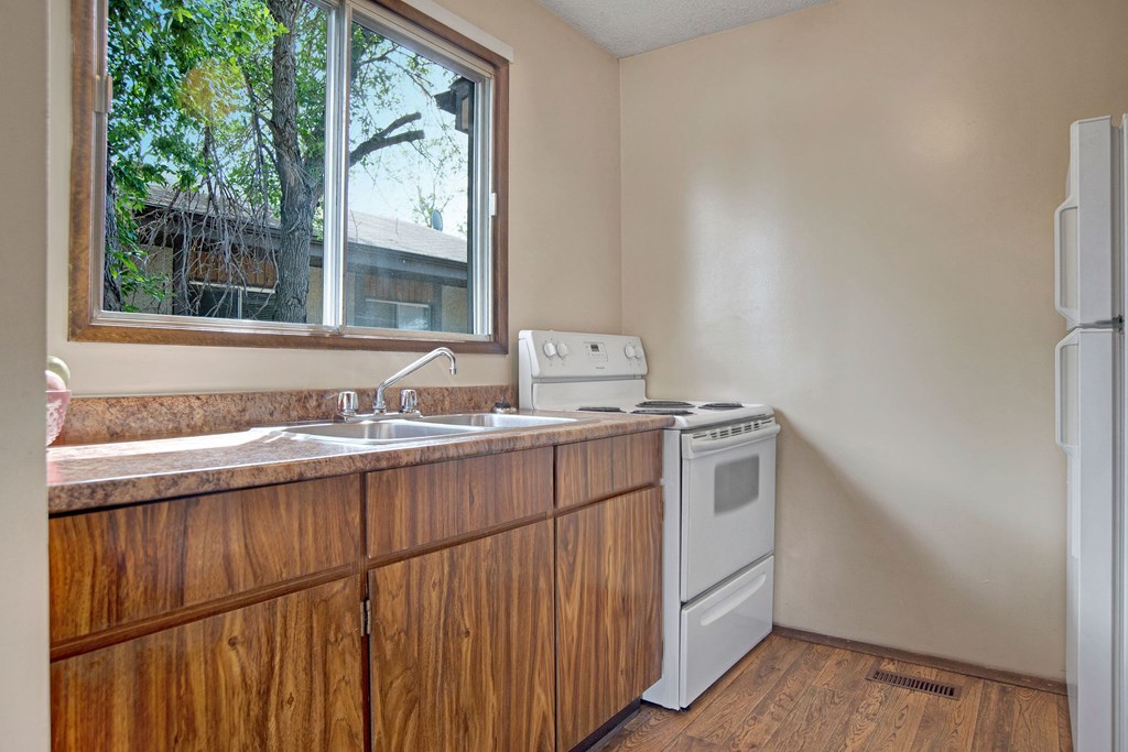 Simple kitchen here at Ventana Apartment Homes with wood cabinets, a white stove, and sink beneath a window showing tree branches outside. Neutral tones, bright daylight.