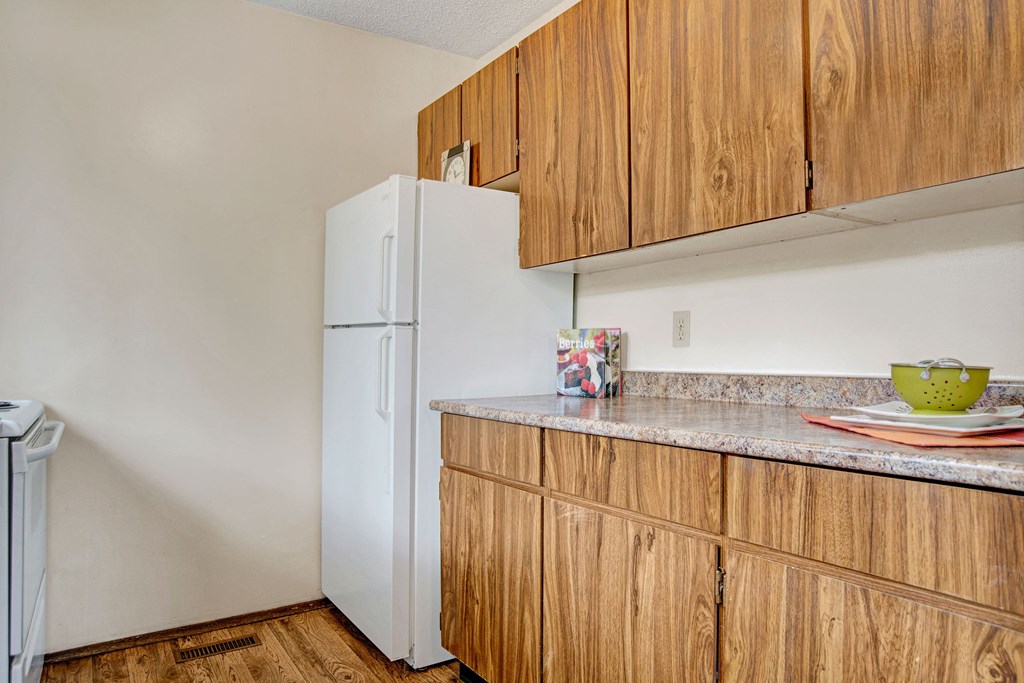 A small kitchen here at Ventana Apartment Homes with wooden cabinets, a white refrigerator, and a countertop displaying a cookbook and a green colander, creating a cozy ambiance.