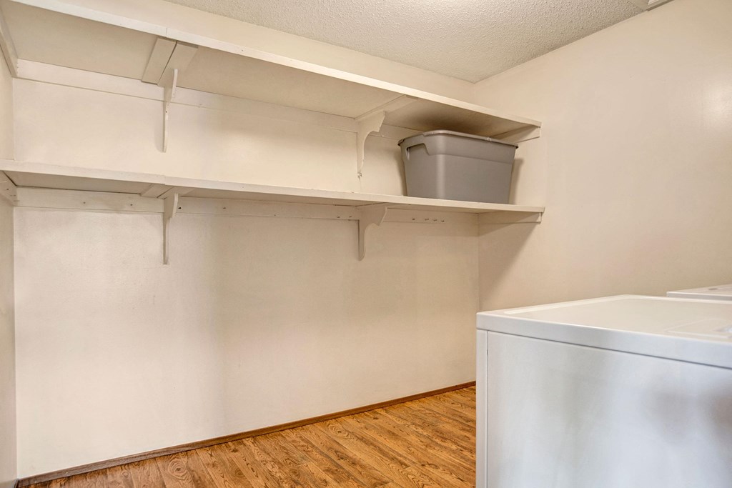Laundry room here at Ventana Apartment Homes with white walls and wooden floor. Two white appliances, likely washer and dryer, are on the right. Shelves above hold a gray storage bin.