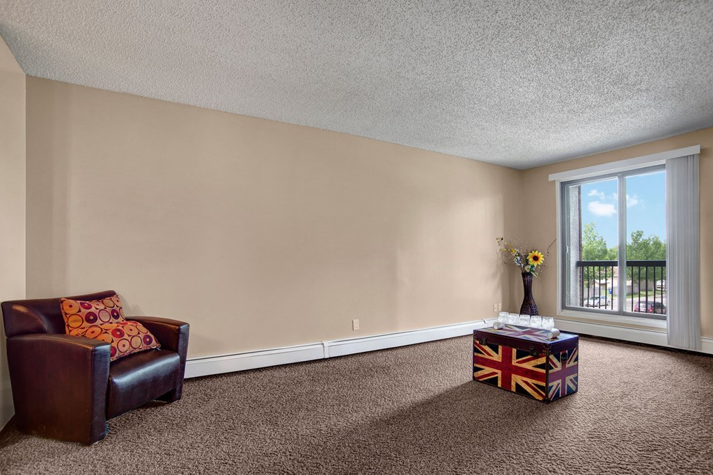 A minimalist room here at Ventana Apartment Homes with a brown armchair and patterned pillow on the left. A Union Jack trunk and a vase with sunflowers sit near a large window on the right.