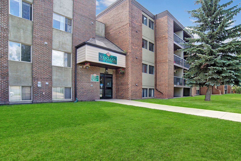 Red brick Ventana Apartment Homes building named Ventana with large windows and a green lawn. A tree and a pathway are in the foreground, under a clear blue sky.
