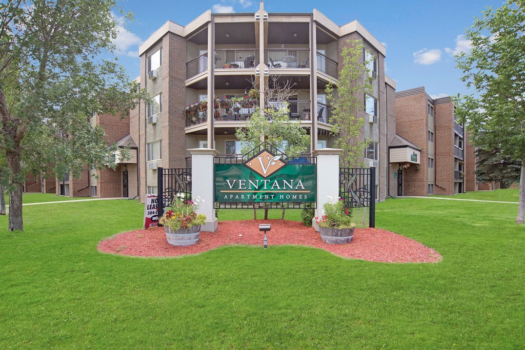 Apartment complex with brick facade, balconies adorned with flowers. A sign reading "Ventana Apartment Homes" is set in a landscaped area. Peaceful ambiance.