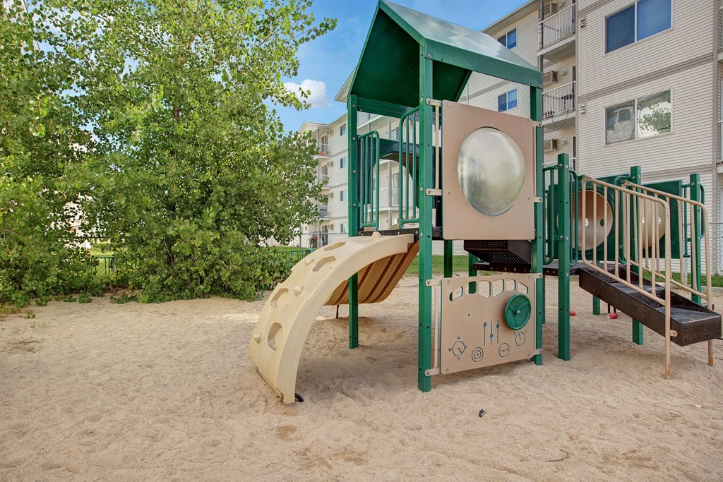 Playground scene here at Villagio Apartment Homes with a green and beige slide set on sandy ground. Lush trees in the background and residential buildings create a serene, inviting atmosphere.