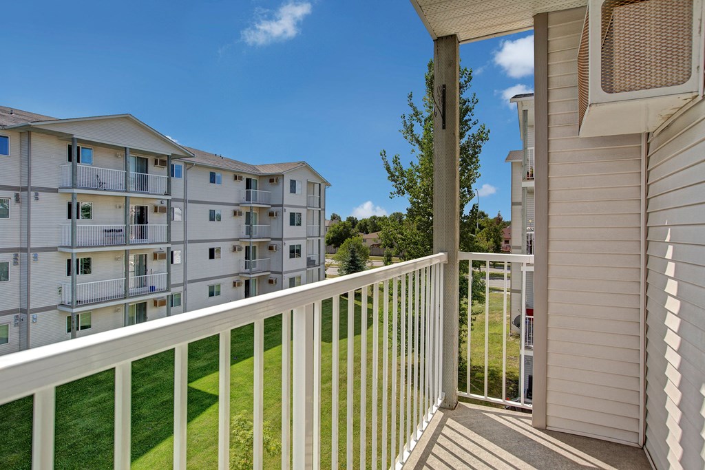 View from a balcony here at Villagio Apartment Homes with white railing, overlooking a grassy area and a multi-story apartment building under a clear blue sky. Peaceful atmosphere.