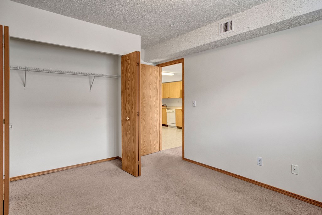 Empty room here at Villagio Apartment Homes with beige carpet, white walls, and an open wooden closet. A door leads to a kitchen with light wood cabinets, creating a neutral, open tone.