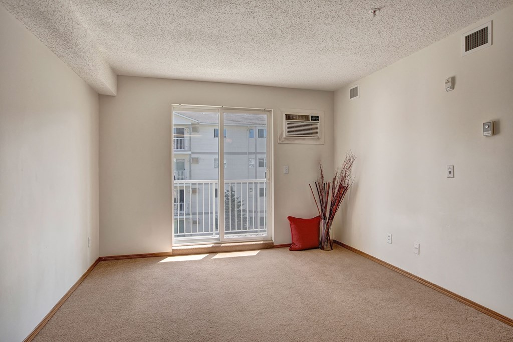 Empty room here at Villagio Apartment Homes with beige carpet, white walls, and a large window letting in natural light. A red cushion and decorative branches add a cozy touch.