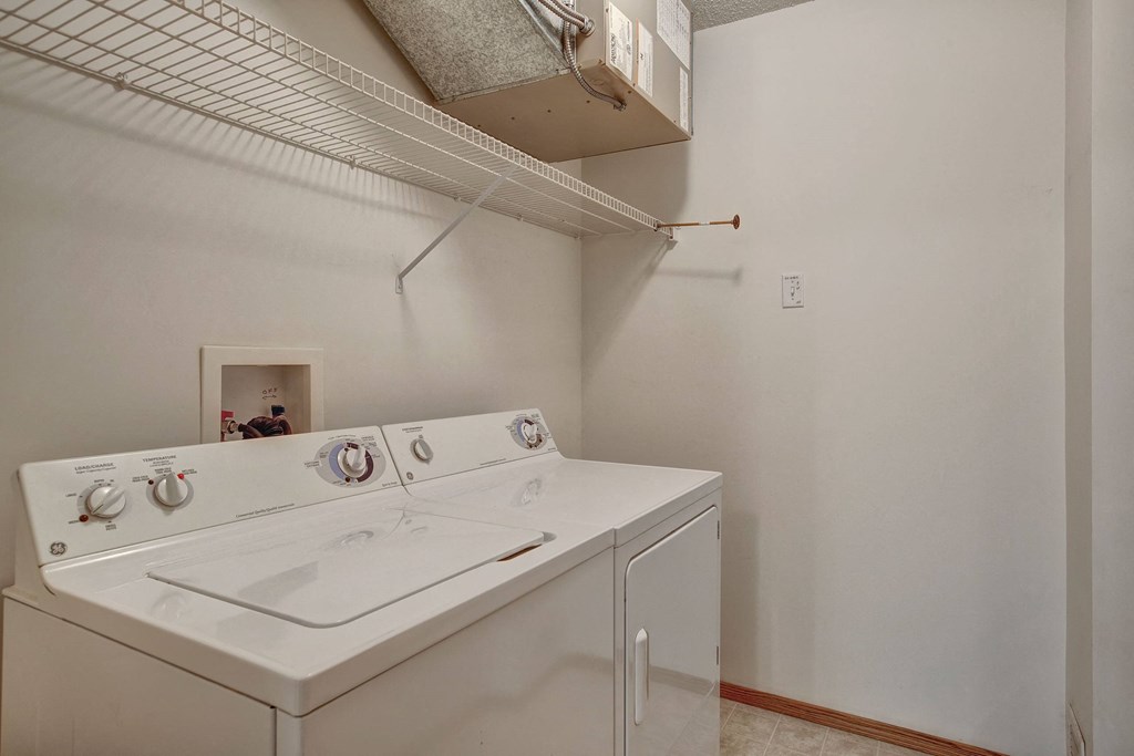 Small laundry room here at Villagio Apartment Homes with a white washer and dryer, white wire shelving above, and neutral walls. The space is clean and organized, conveying simplicity.