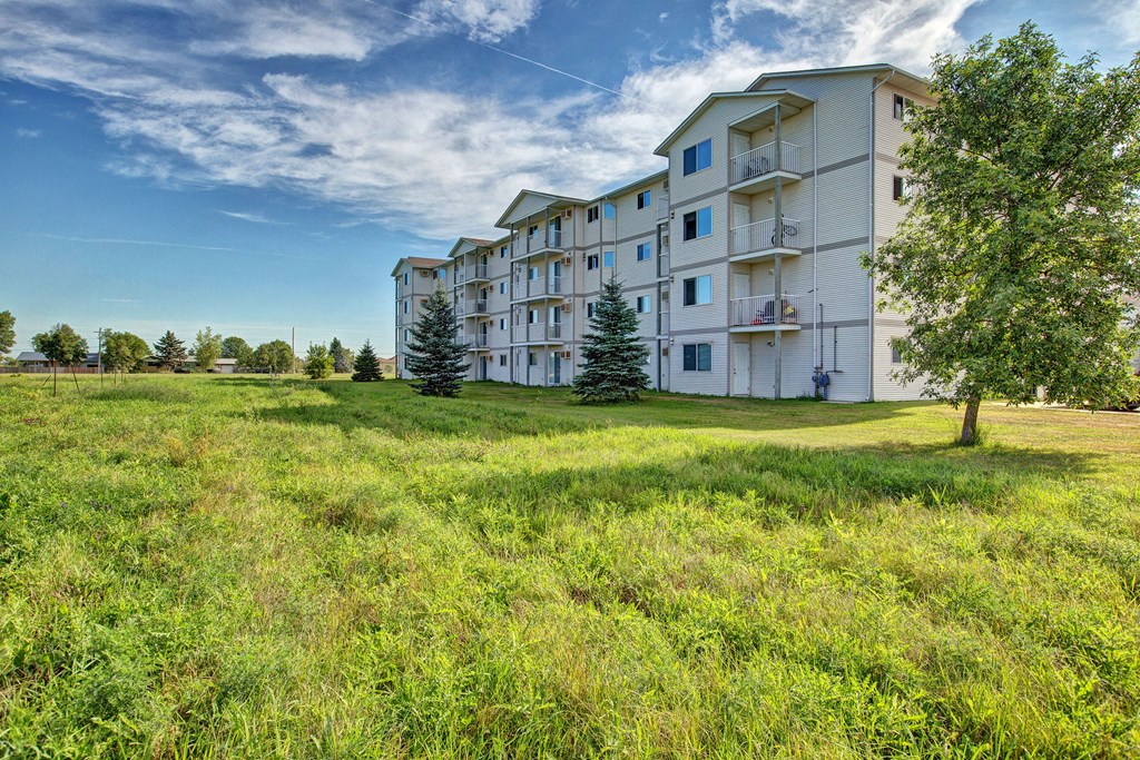 Four-story white Villagio Apartment Homes building with balconies on a sunny day, surrounded by a lush grassy lawn and trees. Blue sky with scattered clouds.
