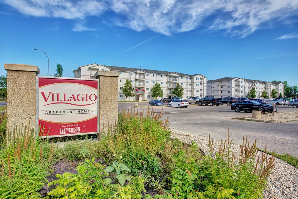 Sign reads "Villagio Apartment Homes" in front of a modern apartment complex with a parking lot and landscaped greenery under a clear blue sky.