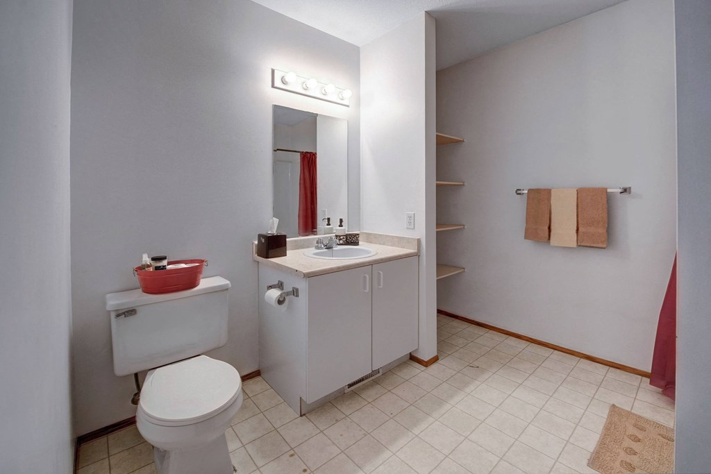 A tidy bathroom here at Willow View Apartment Homes with white walls, a sink with a large mirror above, a toilet topped with red basket, tan towels on a rack, and red shower curtain.