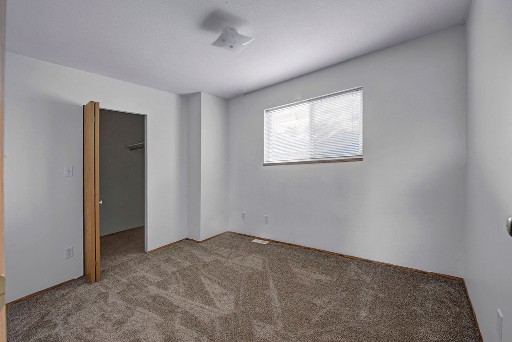 A bedroom with a large walk in closet Small empty room here at Willow View Apartment Homes with beige carpet, white walls, and a window with blinds. An open closet door and ceiling light add to the clean, simple look.
