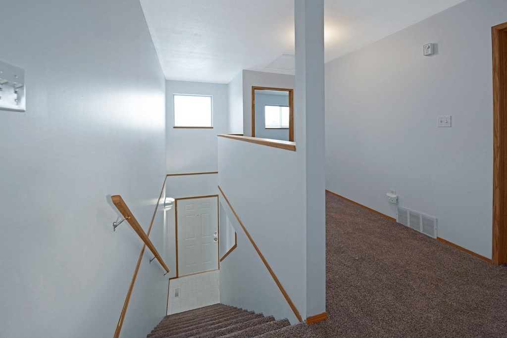 Staircase with wooden handrail in a well-lit hallway here at Willow View Apartment Homes. Carpeted landing leads to a white door. Natural light from a window creates a welcoming atmosphere.