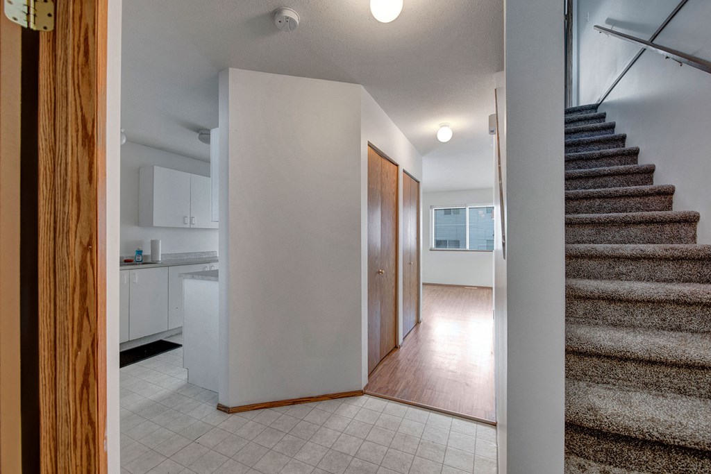 Hallway here at Willow View Apartment Homes with tiled and wood floors, stairs to the right, and a glimpse of a white kitchen. Light filters through a window, creating a cozy ambiance.