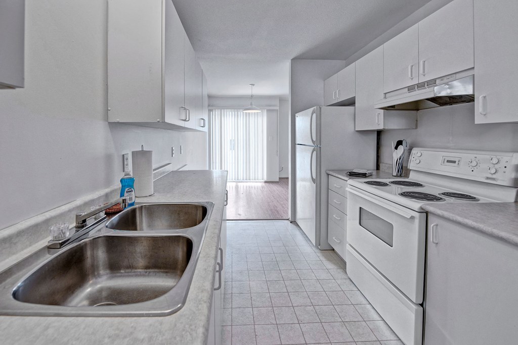 A kitchen with white appliances and white cabinetry A clean, minimalist kitchen here at Willow View Apartment Homes with white cabinets and appliances, including a double sink and stovetop. Sunlight filters through vertical blinds, creating a bright, serene atmosphere.