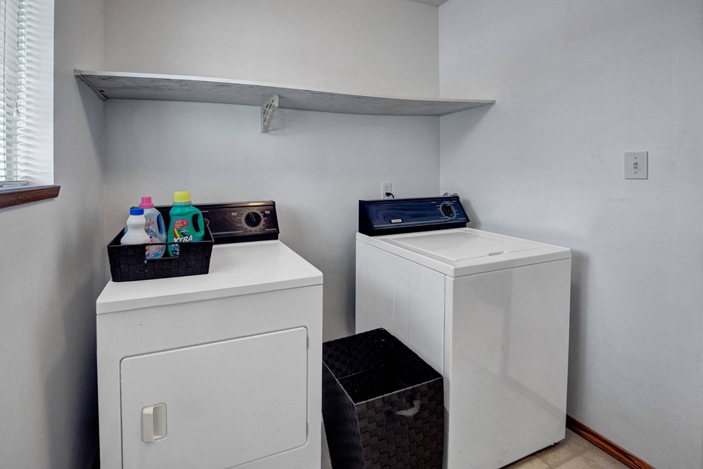 Compact laundry room here at Willow View Apartment Homes with white washer and dryer beneath a white shelf. A basket holds detergent and softener. Neutral tones and neat arrangement.