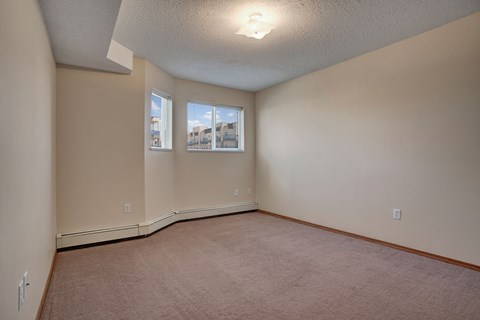 Empty room here at Windsor Terrace Apartment Homes with beige walls and carpet, and two windows showing blue sky. A ceiling light illuminates the space with a calming ambience.