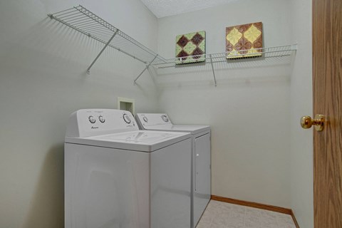 Laundry room here at Windsor Terrace Apartment Homes with a white washer and dryer side by side under a wall-mounted wire shelf. Two decorative tiles hang above the appliances. Clean and organized.