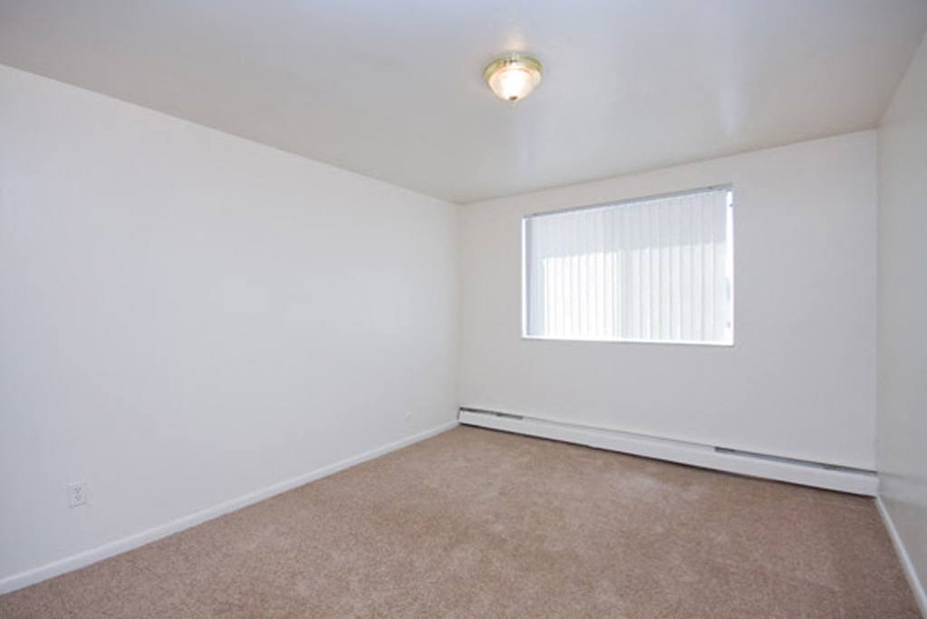 Bristol Square Bedroom Empty room here at Bristol Square Apartment Homes with white walls, beige carpet, and a ceiling light fixture. A window with vertical blinds allows natural light, creating a neutral, calm tone.