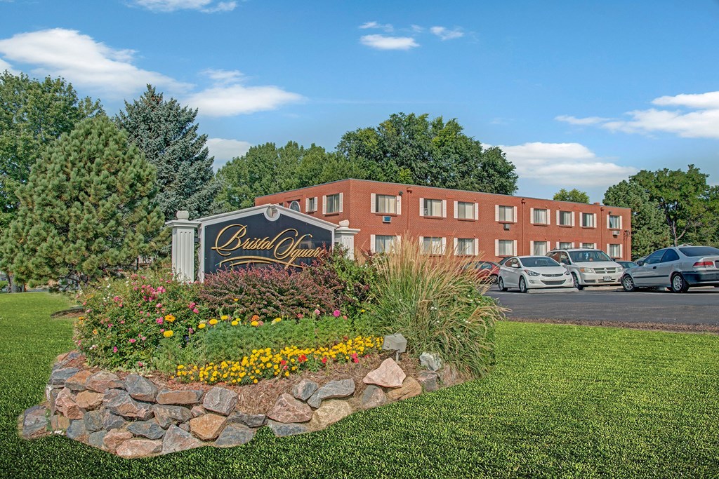 Bristol Square Building & Sign Manicured garden with colorful flowers and a "Bristol Square" sign in front. A brick residential building with parked cars is visible behind, under clear blue sky.