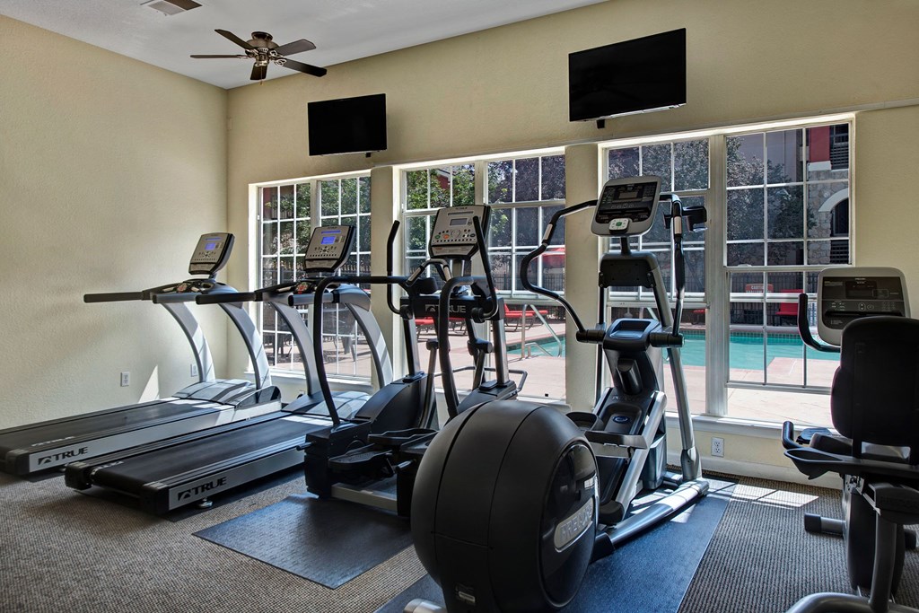 Camelback Pointe Cardio Equipment Facing Pool A well-lit gym room here at Camelback Pointe Apartment Homes with two treadmills and two elliptical machines facing large windows overlooking a pool area. Monitors and a fan are visible above.