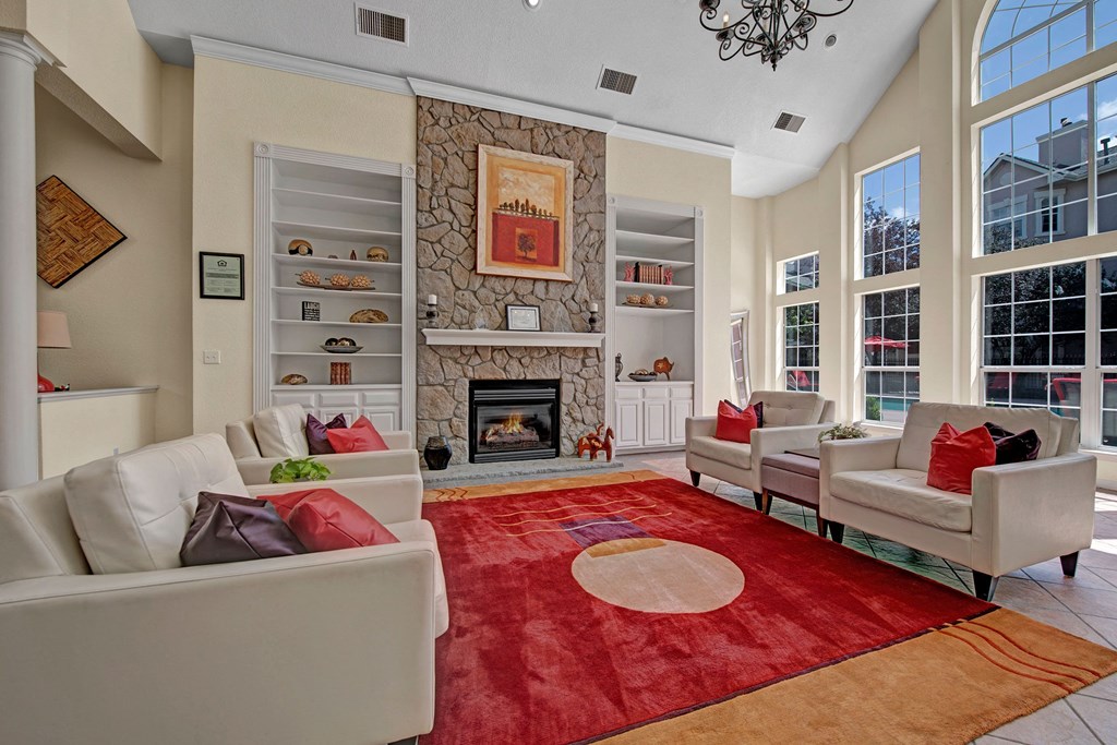 Bright, spacious resident lounge here at Camelback Pointe Apartment Homes with high ceilings, a stone fireplace, white shelves, and large windows. Red and beige carpet with cream sofas.