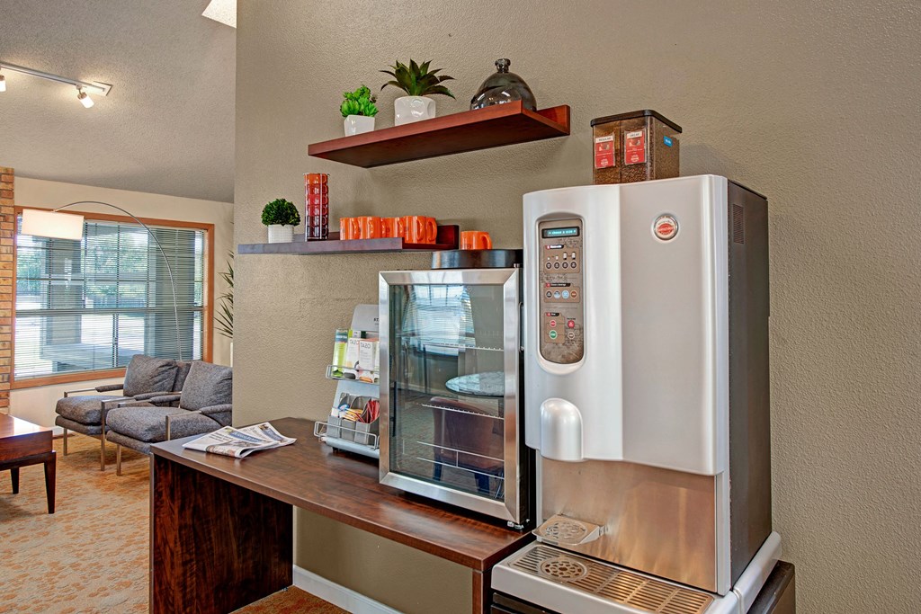 Grand View Resident Coffee Machine A modern coffee station in an office lounge here at Grand View Apartment Homes with a coffee machine on a wooden counter, a small snack display, and wall shelves holding plants and mugs.