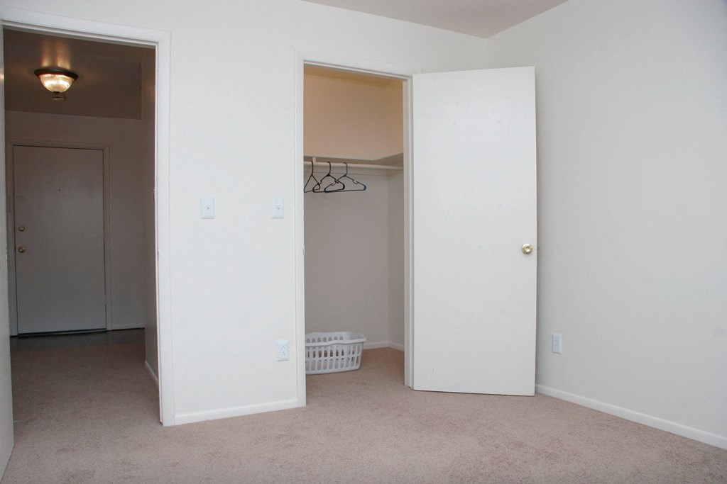 Hyde Park North Bedroom & Closet Empty room here at Hyde Park North Apartment Homes with beige carpet, white walls, and an open closet door showing hangers and a laundry basket. The room has a simple, clean appearance.