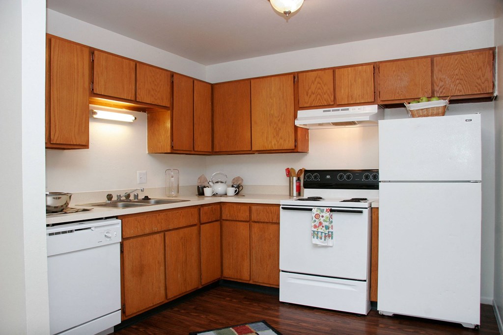 Hyde Park North Kitchen Appliances Compact kitchen here at Hyde Park North Apartment Homes with wooden cabinets, white appliances, and dark flooring. Includes a stove, fridge, and dishwasher. Bright lighting creates a cozy feel.