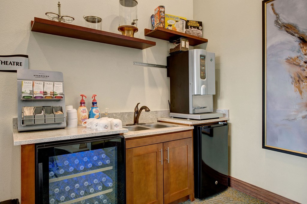 Cozy kitchenette here at La Bella Vita Apartment Homes with a coffee station. Features include a Tazo tea rack, syrup bottles, mugs, water cooler, and brewing machine. Warm, inviting atmosphere.