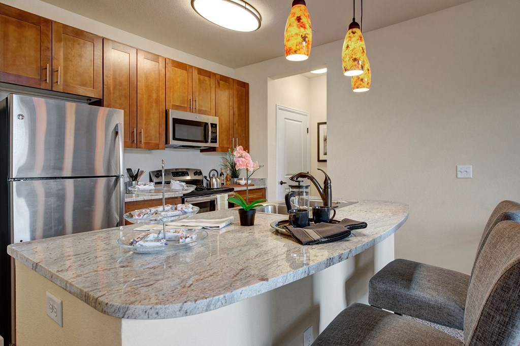 Elegant kitchen here at La Bella Vita Apartment Homes with wooden cabinets, granite countertop, and stainless steel appliances. Warm lighting from orange pendant lights and a cozy bar area with stools.
