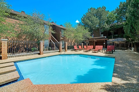 Windtree Apartments Pool Sunny day at a serene outdoor pool area here at Windtree Apartment Homes surrounded by trees and brick buildings. Several cushioned lounge chairs lined up, creating a relaxing atmosphere.