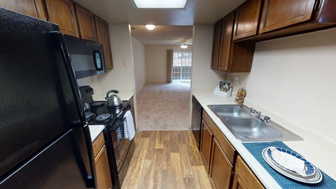 Windtree Apartments Kitchen Compact kitchen here at Windtree Apartment Homes with a black stove, microwave, and fridge on the left. Brown cabinets above. Beige counters hold a double sink and kettle. Wooden floor leads to a bright living room with carpet, glass door, and ceiling fan. Cozy and inviting atmosphere.