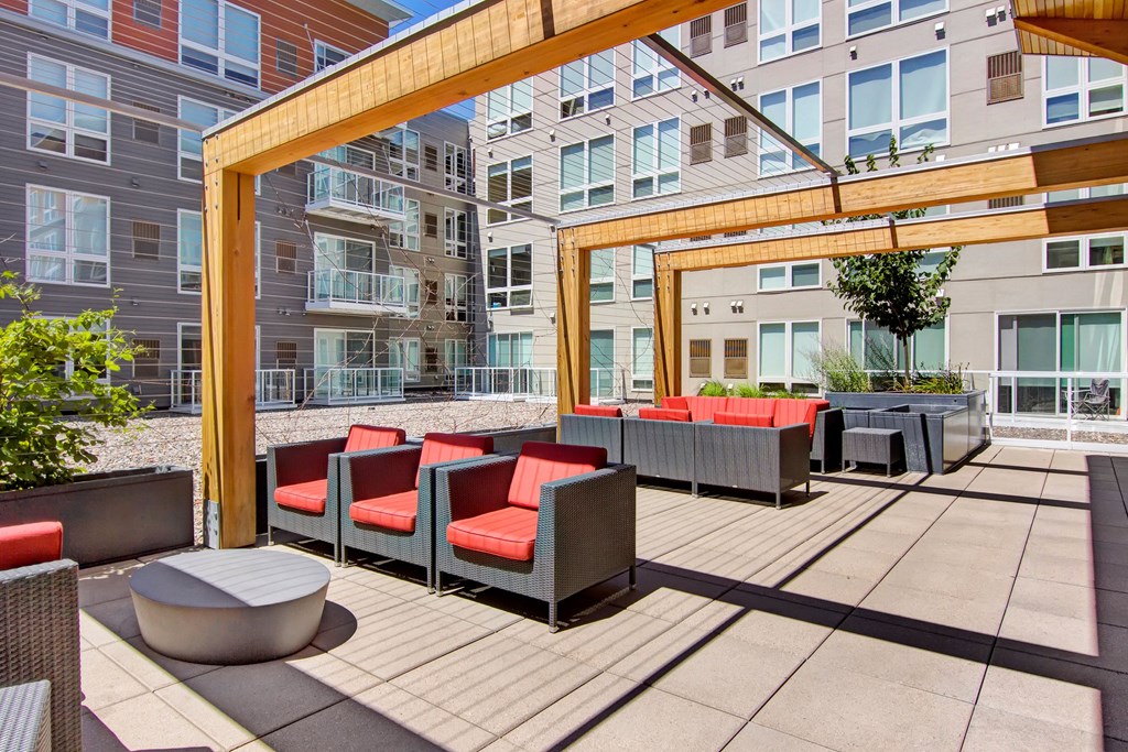 Modern apartment courtyard here at 7west Apartment Homes with gray buildings, featuring wooden pergolas and red-cushioned patio furniture. Bright, sunny atmosphere with a relaxed vibe.