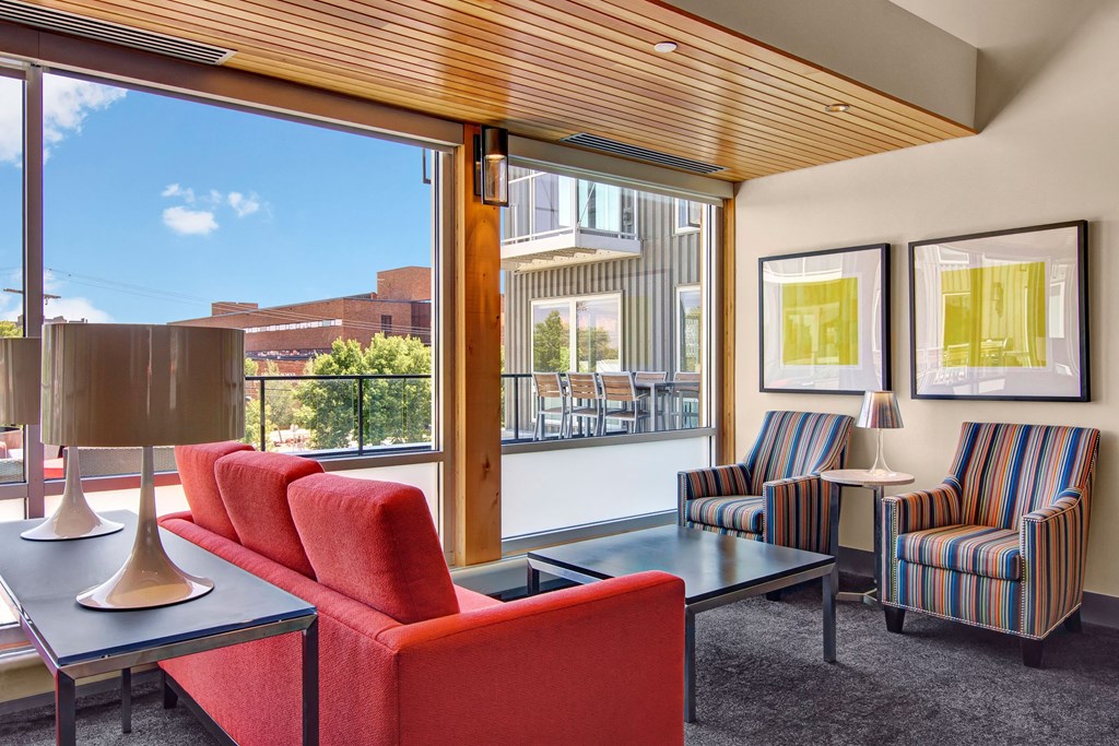 Modern lounge here at 7west Apartment Homes with a red sofa and striped armchairs near large windows. Bright, airy space features abstract art and a patio view. Cozy ambiance.