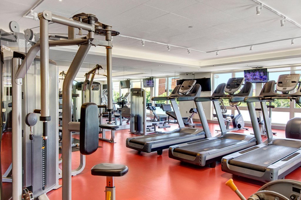 Modern gym here at 7west Apartment Homes with red flooring, featuring treadmills, weight machines, and large windows allowing natural light. Energetic and inviting atmosphere.