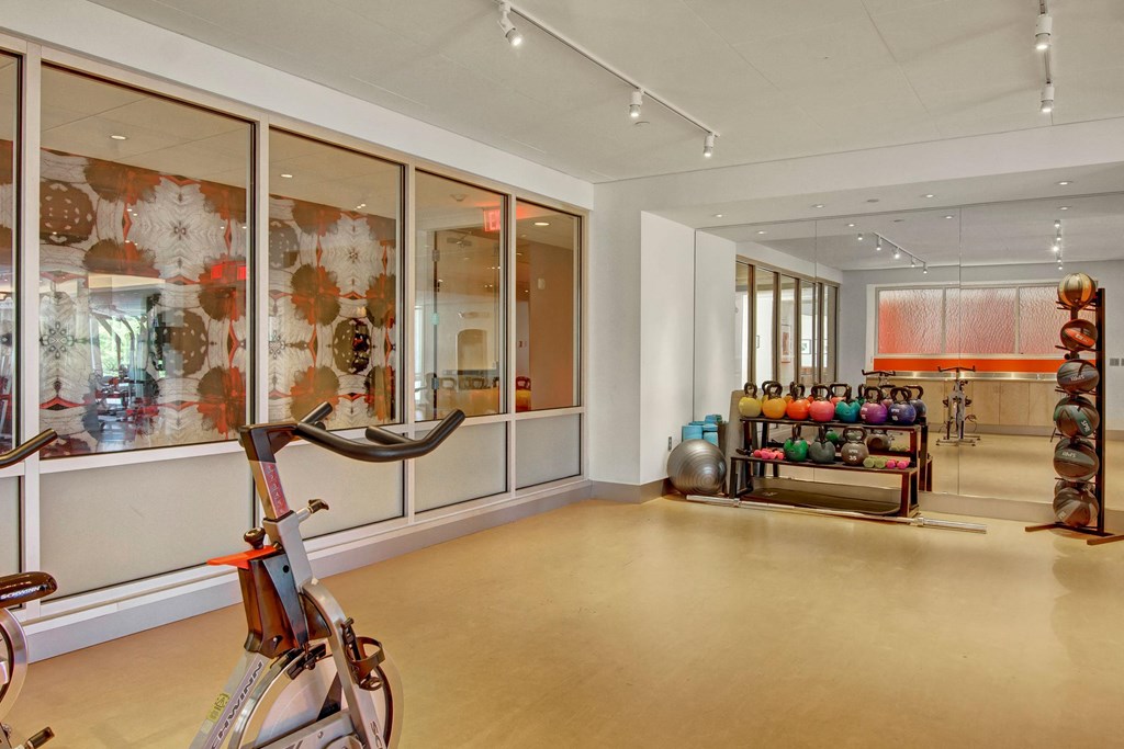 Bright gym room here at 7west Apartment Homes with exercise bikes, colorful kettlebells, medicine balls, and a stability ball. Modern decor includes abstract art on glass walls.