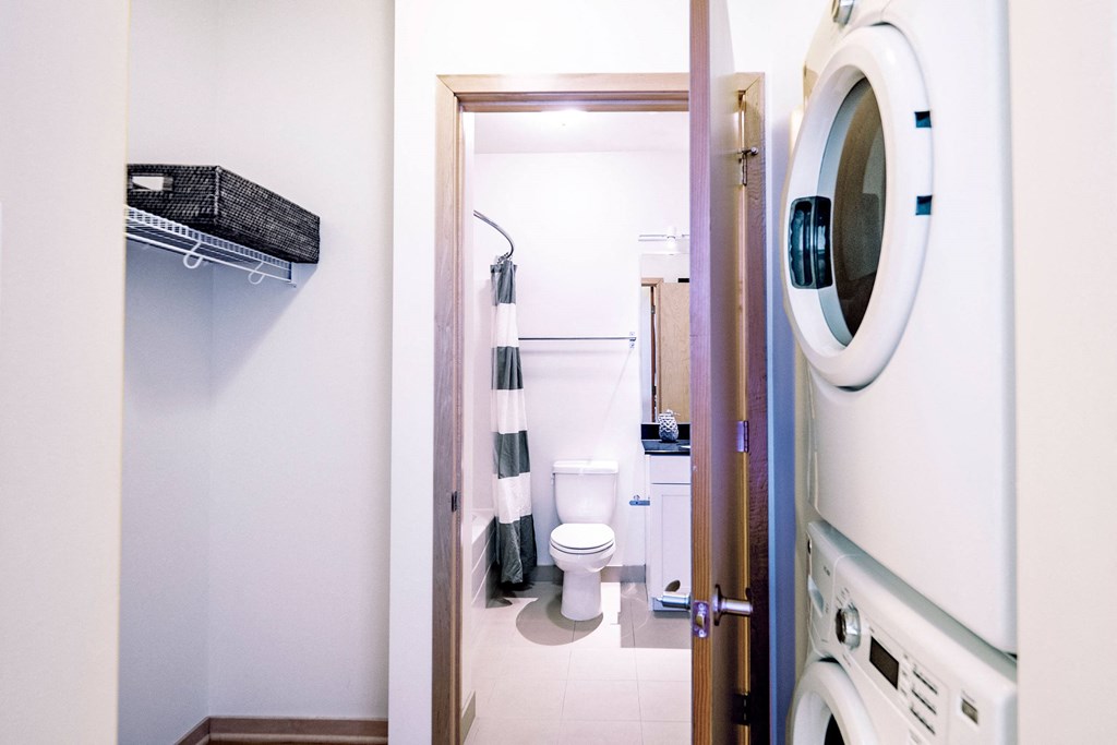 Laundry room here at 7west Apartment Homes with a stacked washer and dryer on the right, and a door opening into a bathroom with a toilet, sink, and striped shower curtain in neutral tones.