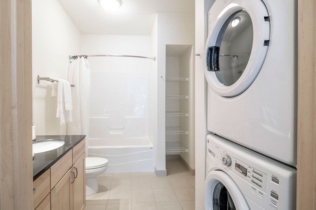 Bright bathroom here at 7west Apartment Homes with a bathtub, toilet, and wooden vanity. A stacked washer and dryer are on the right. Shelving is visible in the wall niche. Minimalist and clean.