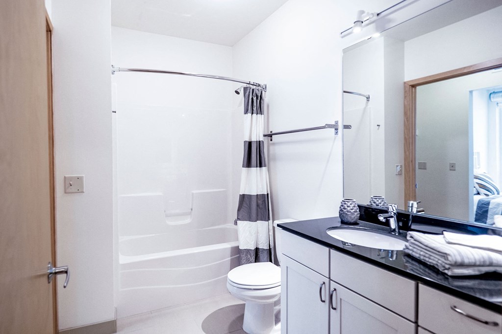 Bright, modern bathroom here at 7west Apartment Homes with white walls, a bathtub with a striped curtain, and a black countertop sink. Clean, minimalist design conveys tranquility.